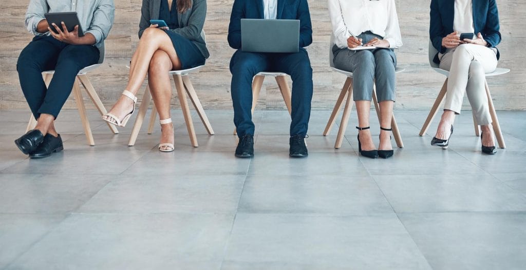 Five business professionals seated in a row, using laptops, tablets, phones, and a notebook while waiting for job interviews.